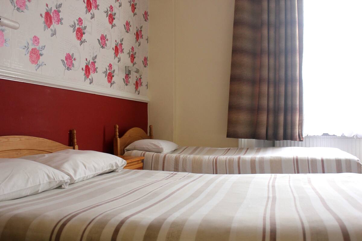 A hotel room with two single beds, a wooden headboard, a floral wallpaper design, and a window with curtains.