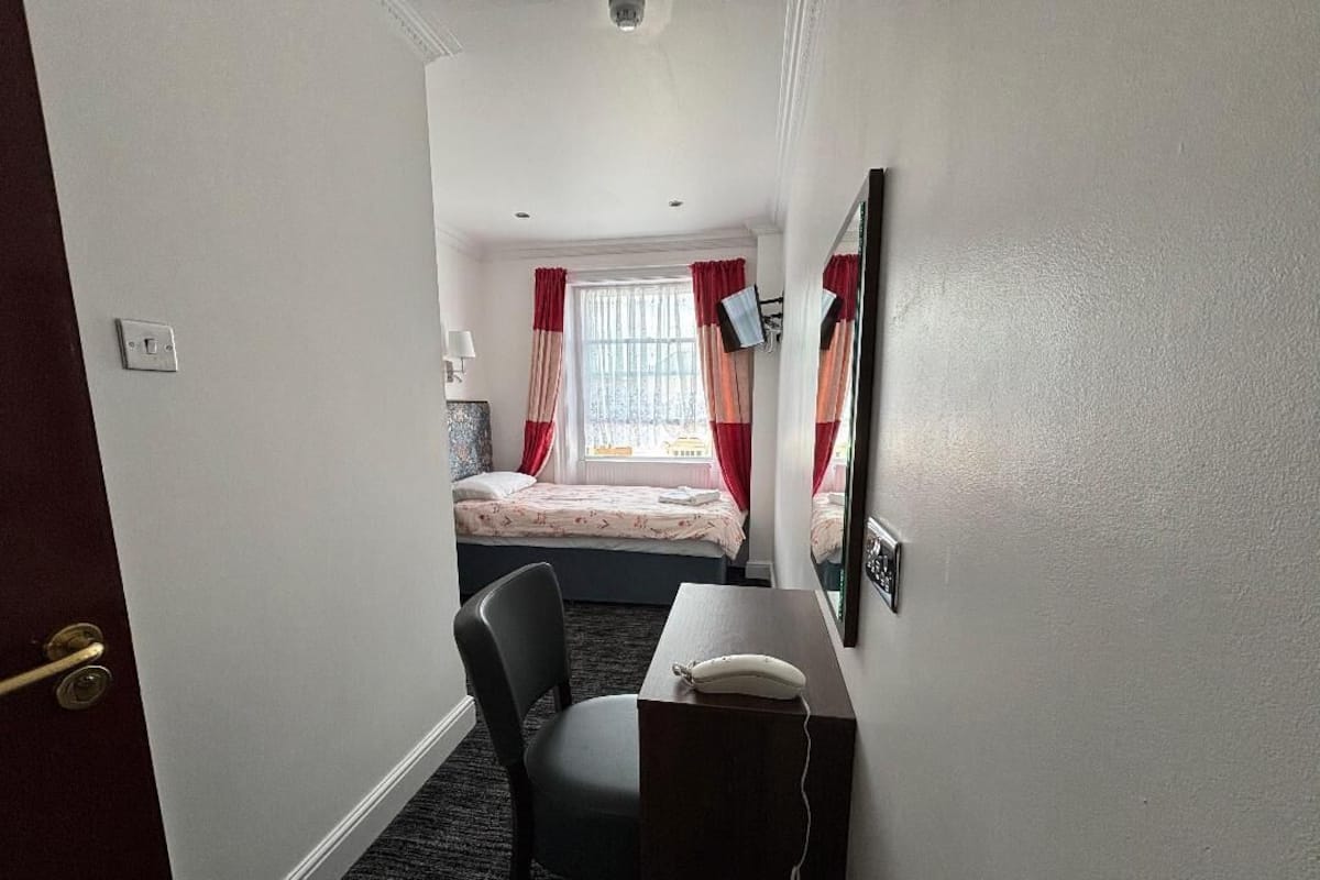 A hotel room with a bed, a desk with a telephone, a chair, a window with curtains, and a mirror on the wall.