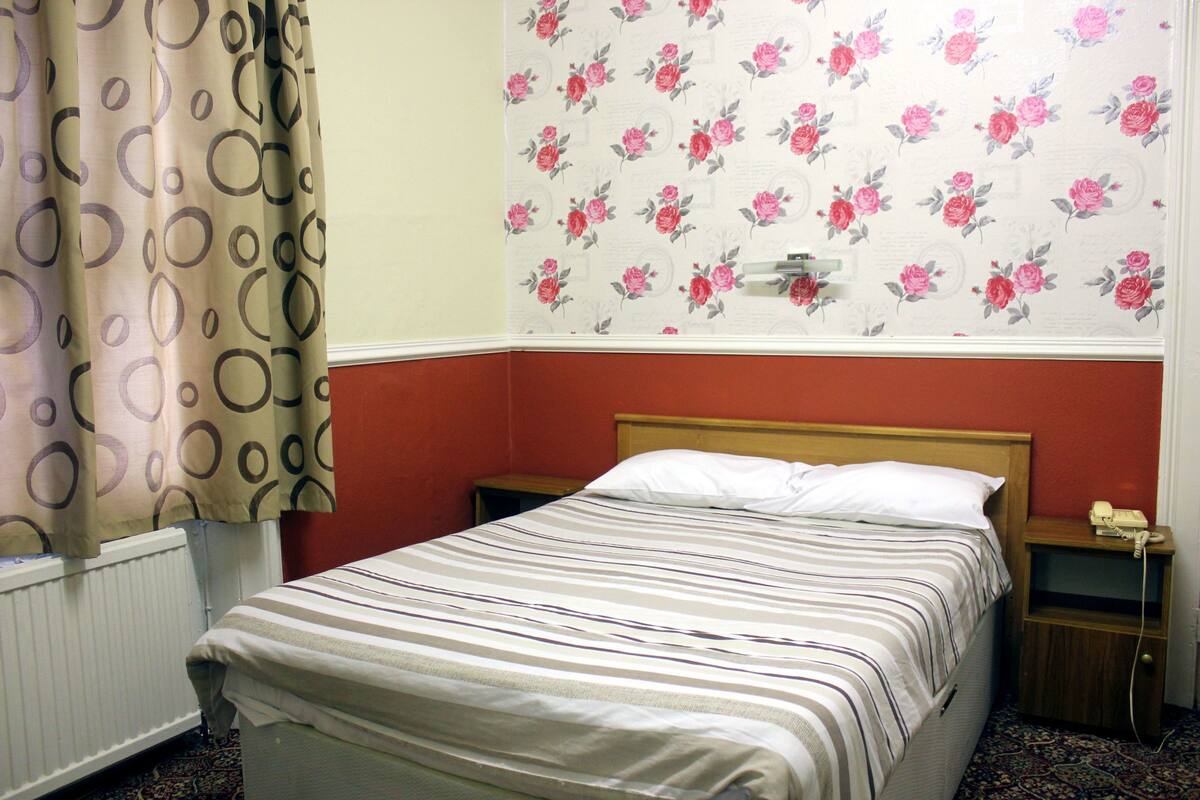 A hotel room with a bed, a nightstand, a radiator, and floral wallpaper.