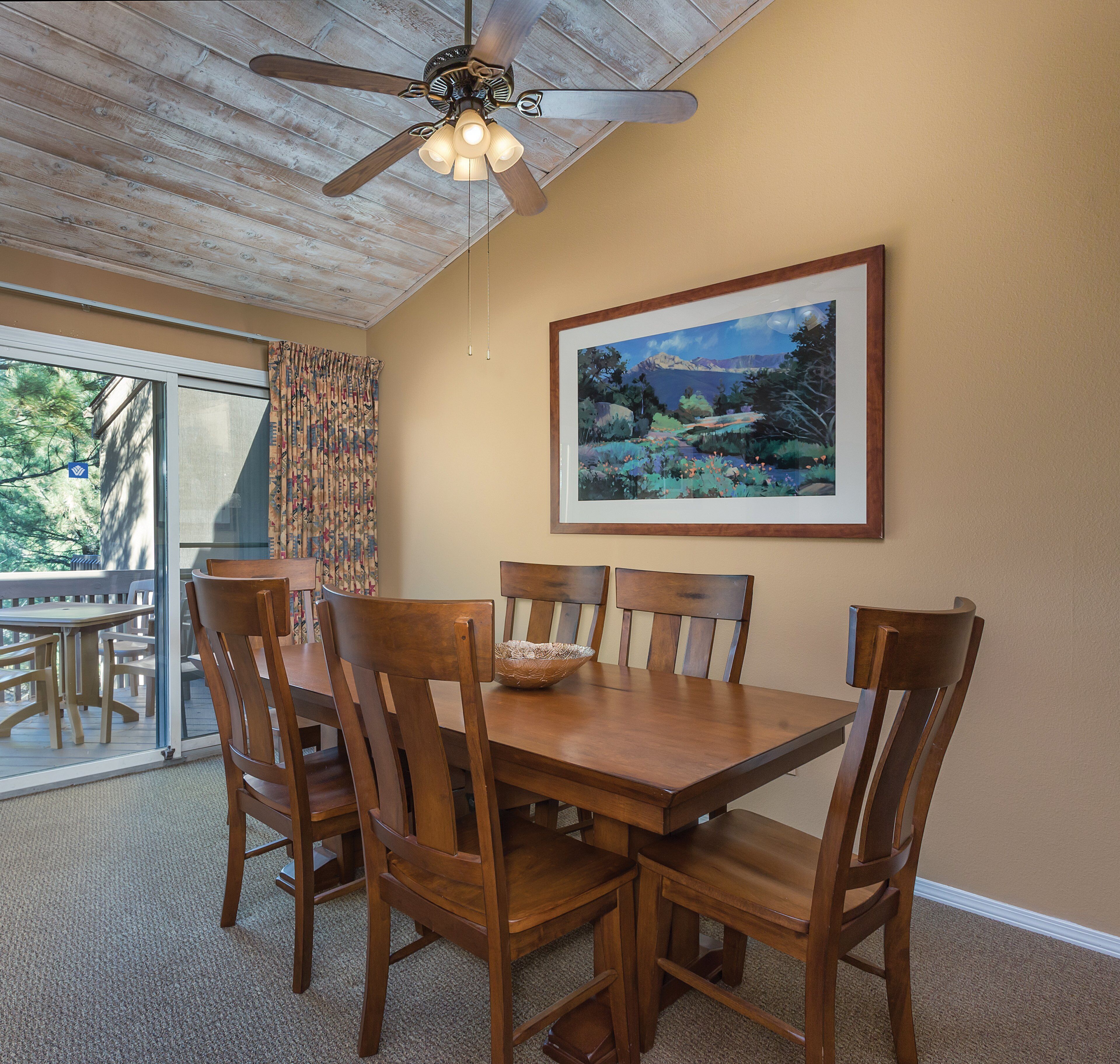 A dining area with a wooden table and chairs, a ceiling fan, and a framed landscape painting on the wall.