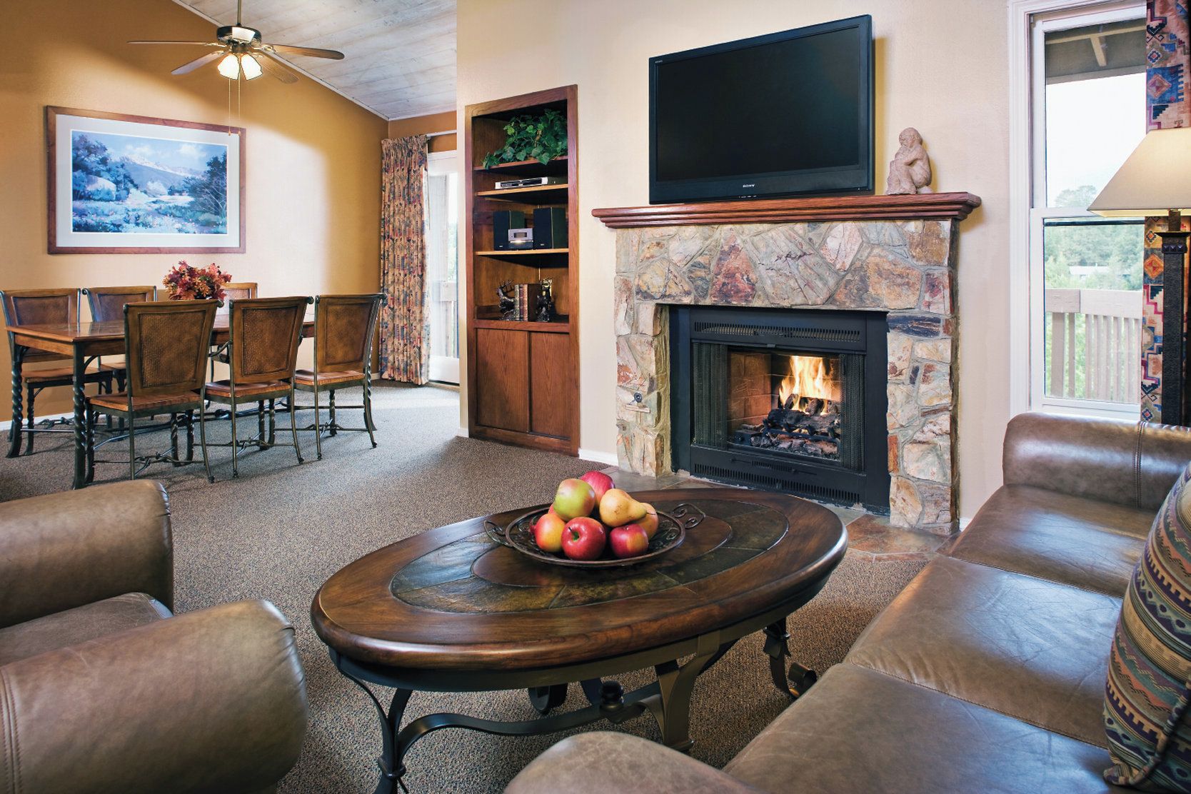 Una sala de estar con chimenea, sofás de cuero, una mesa de centro con frutas, un televisor y un comedor con sillas y una mesa.