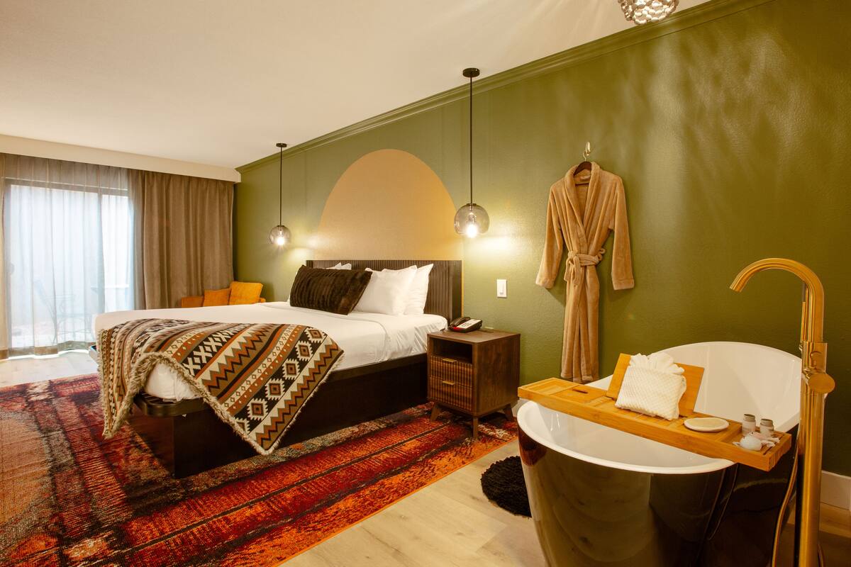 A hotel room with a bed, bedside table, nightstand, and a large bathtub with a wooden tray.