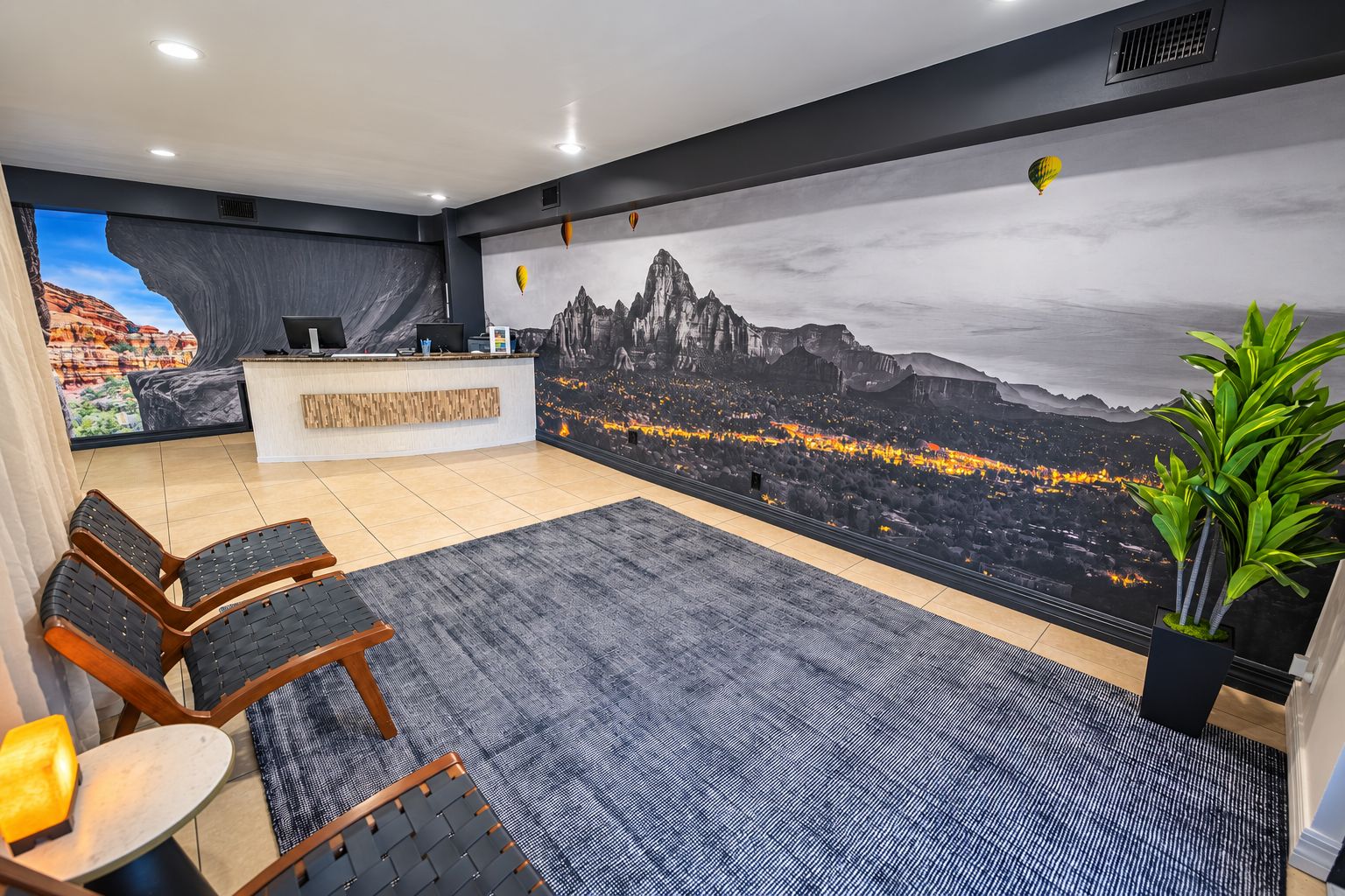 Zona de recepción de un hotel con un gran mural de la ciudad, un mostrador de recepción, un área de descanso y una planta en maceta.