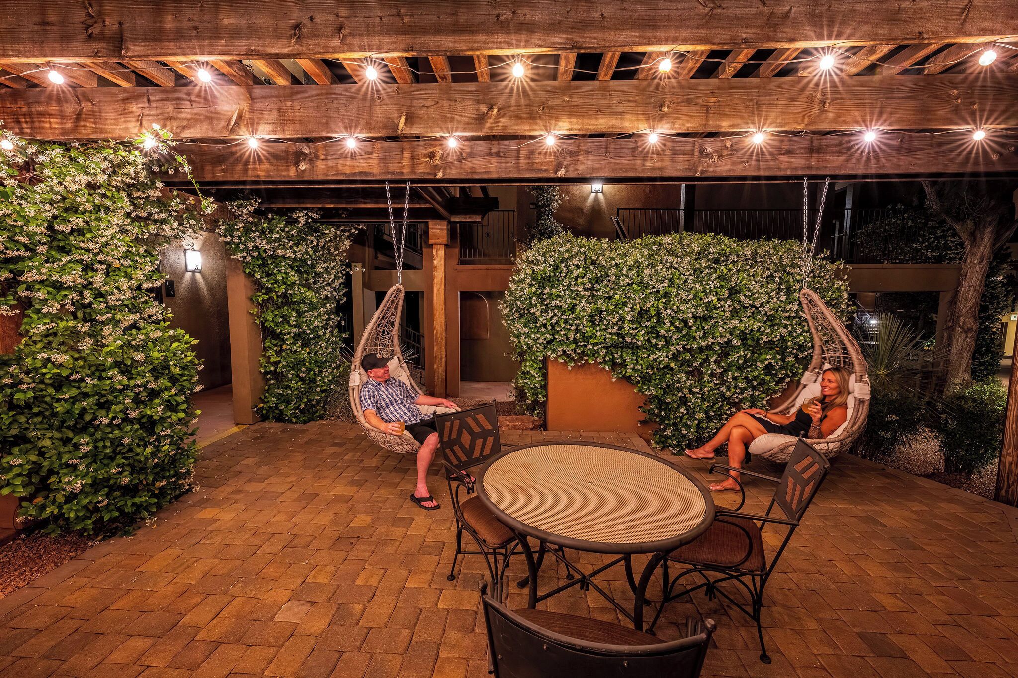 An outdoor patio with wicker chairs and a round table, surrounded by greenery and wooden beams.