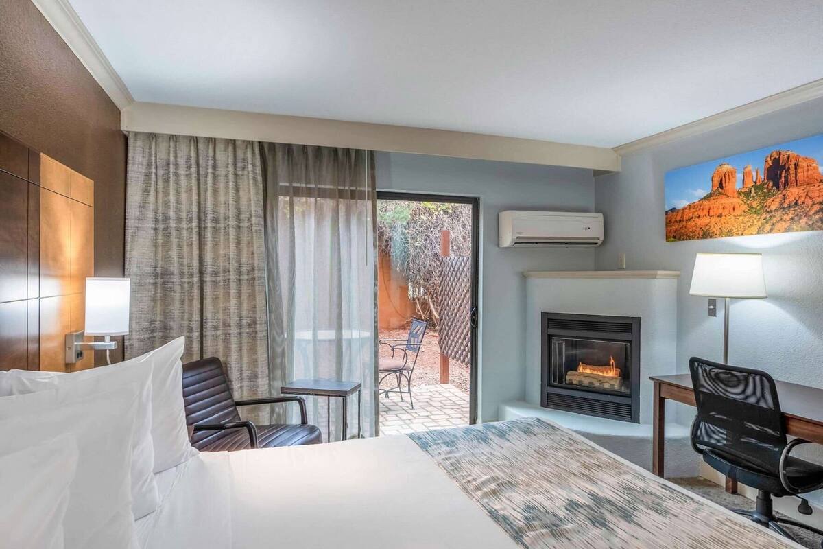 A hotel room with a bed, a desk, a chair, a fireplace, and a view of the outdoors.