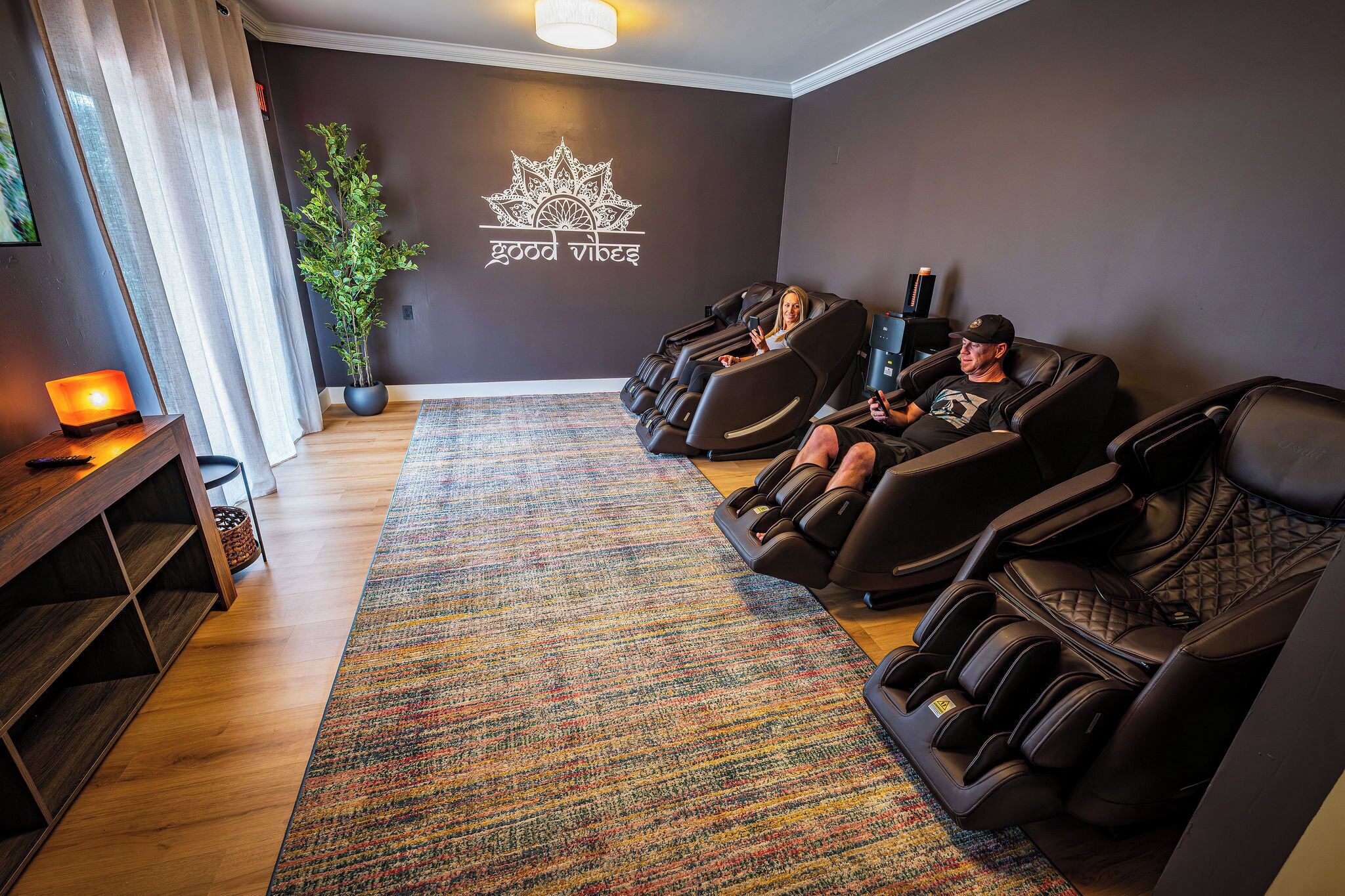Une salle de massage équipée de deux fauteuils de massage, d’une plante en pot, d’un mur orné d’un logo et d’une étagère en bois.