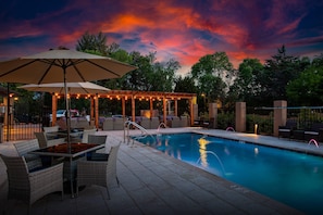 A pool area with lounge chairs and an umbrella, surrounded by a pergola with string lights.