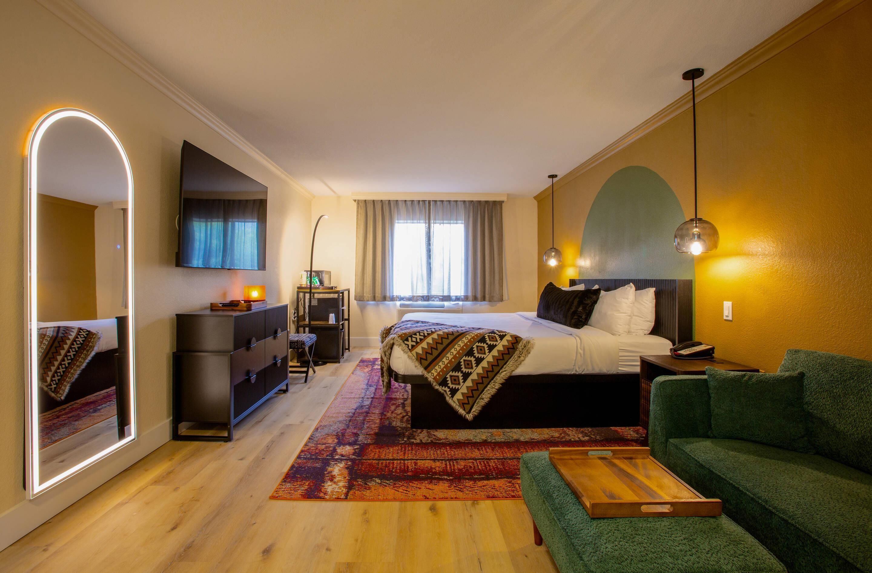 A hotel room with a bed, a green sofa, a desk, a mirror, and a window with curtains.