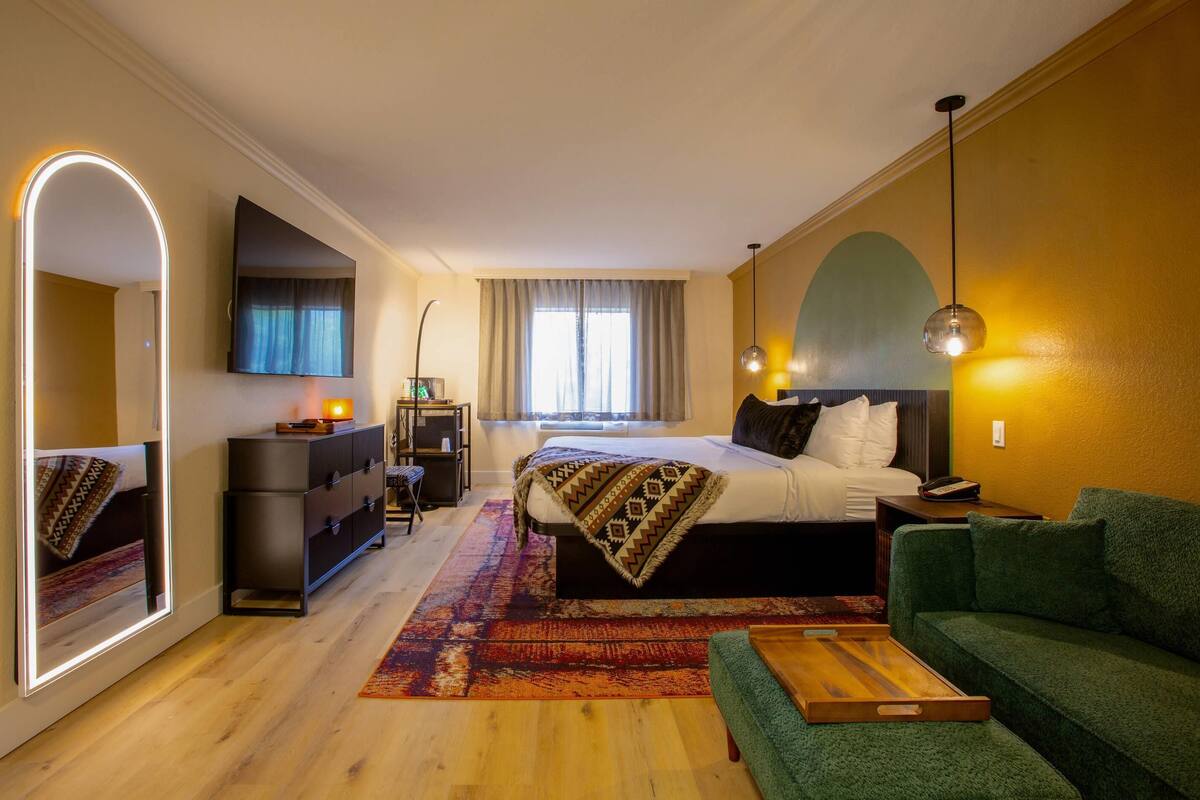 A hotel room with a bed, a green sofa, a desk, a mirror, and a window with curtains.