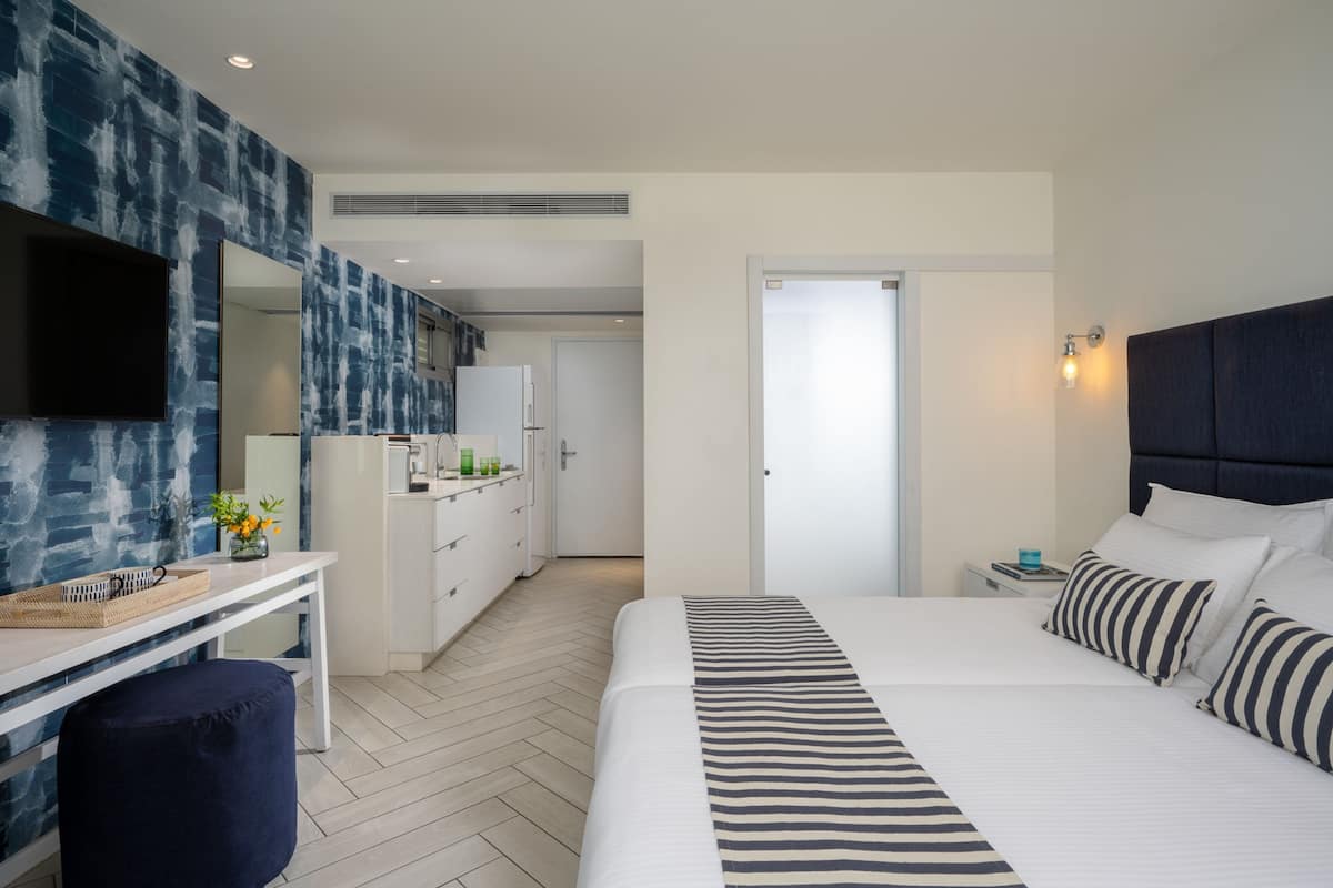 A modern hotel room with a large bed, a desk with a mirror, and a kitchenette in the background.