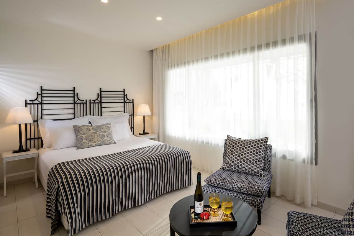 A hotel room with a bed, two bedside tables with lamps, a chair, a small table with a bottle and glasses, and a large window with sheer curtains.