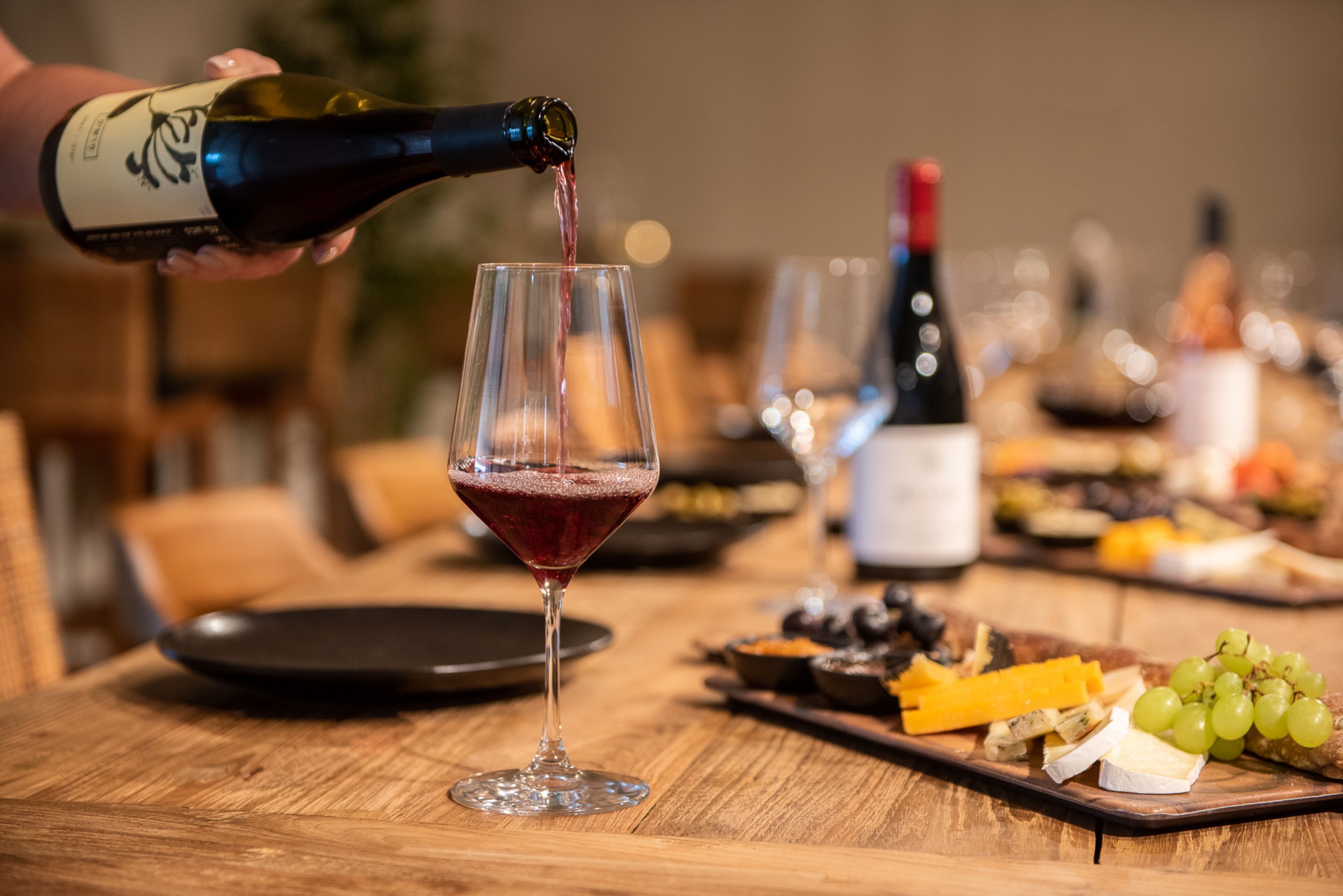 A wine bottle being poured into a glass, a cheese board with various cheeses and grapes, and a bottle of red wine in the background.