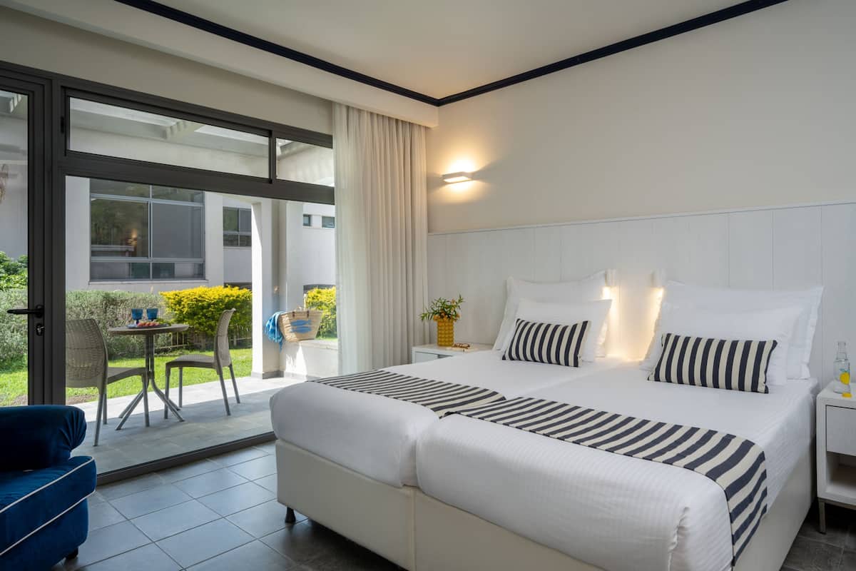 A modern hotel room with a large bed, a small table with chairs, and a view of the outdoors through a sliding glass door.