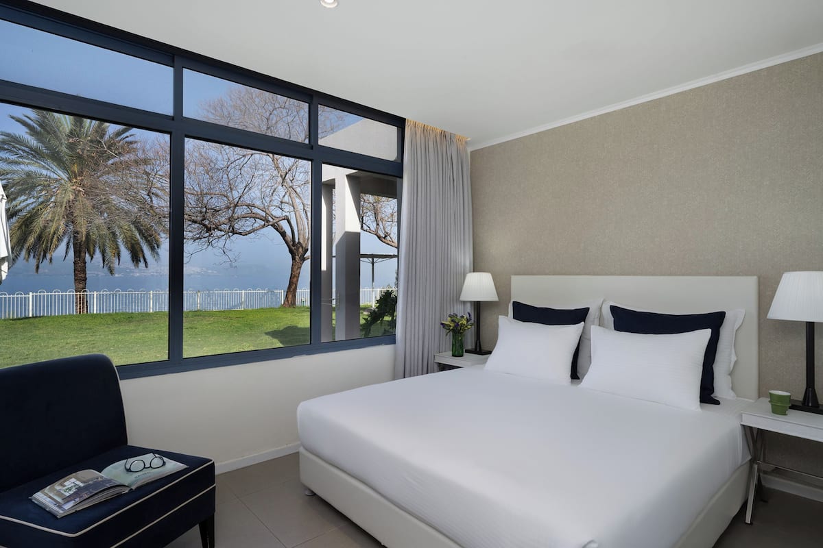 A hotel room with a large bed, a bedside table with a lamp, a chair with a magazine, and a view of the sea and palm trees through large windows.