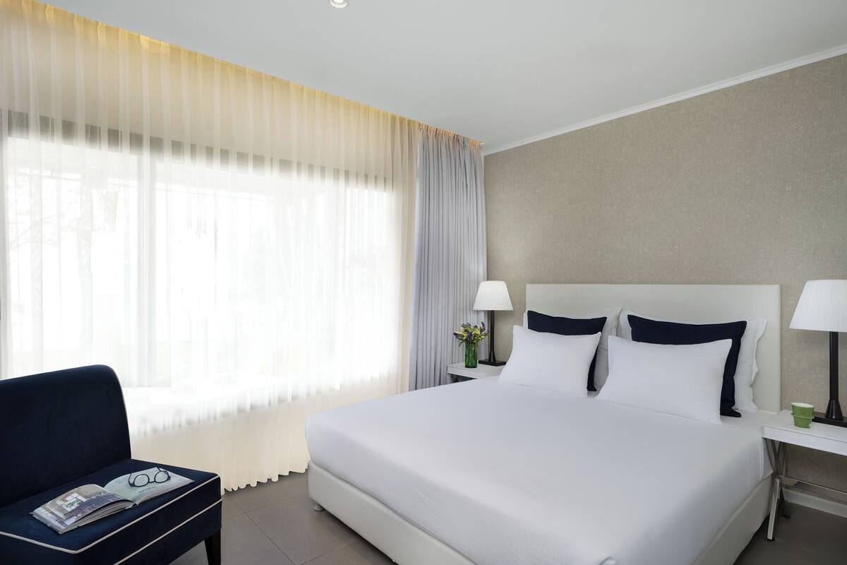 A hotel room with a large bed, two bedside tables, a chair, and a window with sheer curtains.