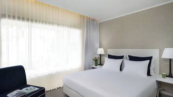 Una habitación de hotel con una cama grande, dos mesitas de noche, una silla y una ventana con cortinas translúcidas.