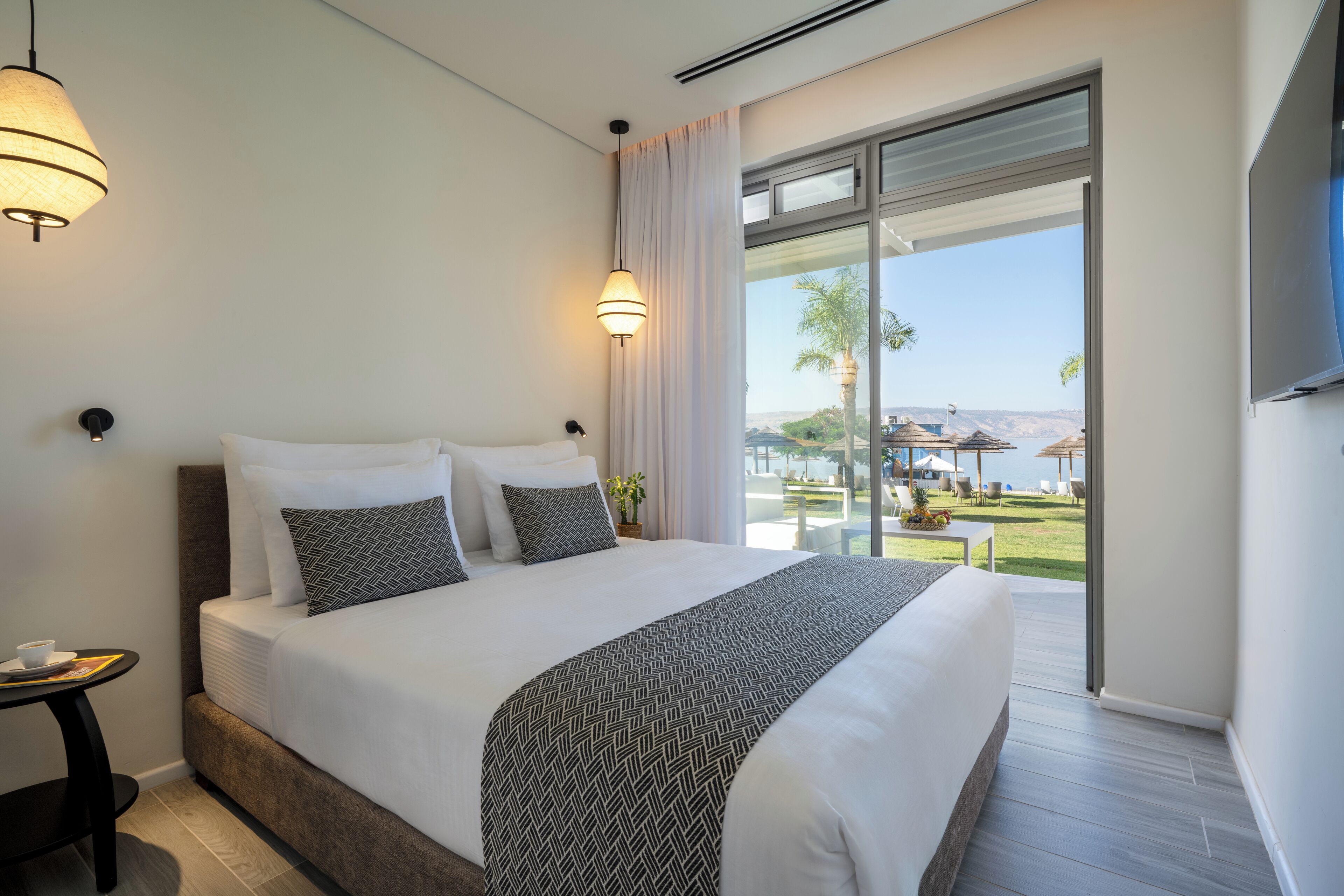 Habitación de hotel moderna con una cama grande, dos lámparas de noche, una mesita con una taza y vistas a la piscina y palmeras.
