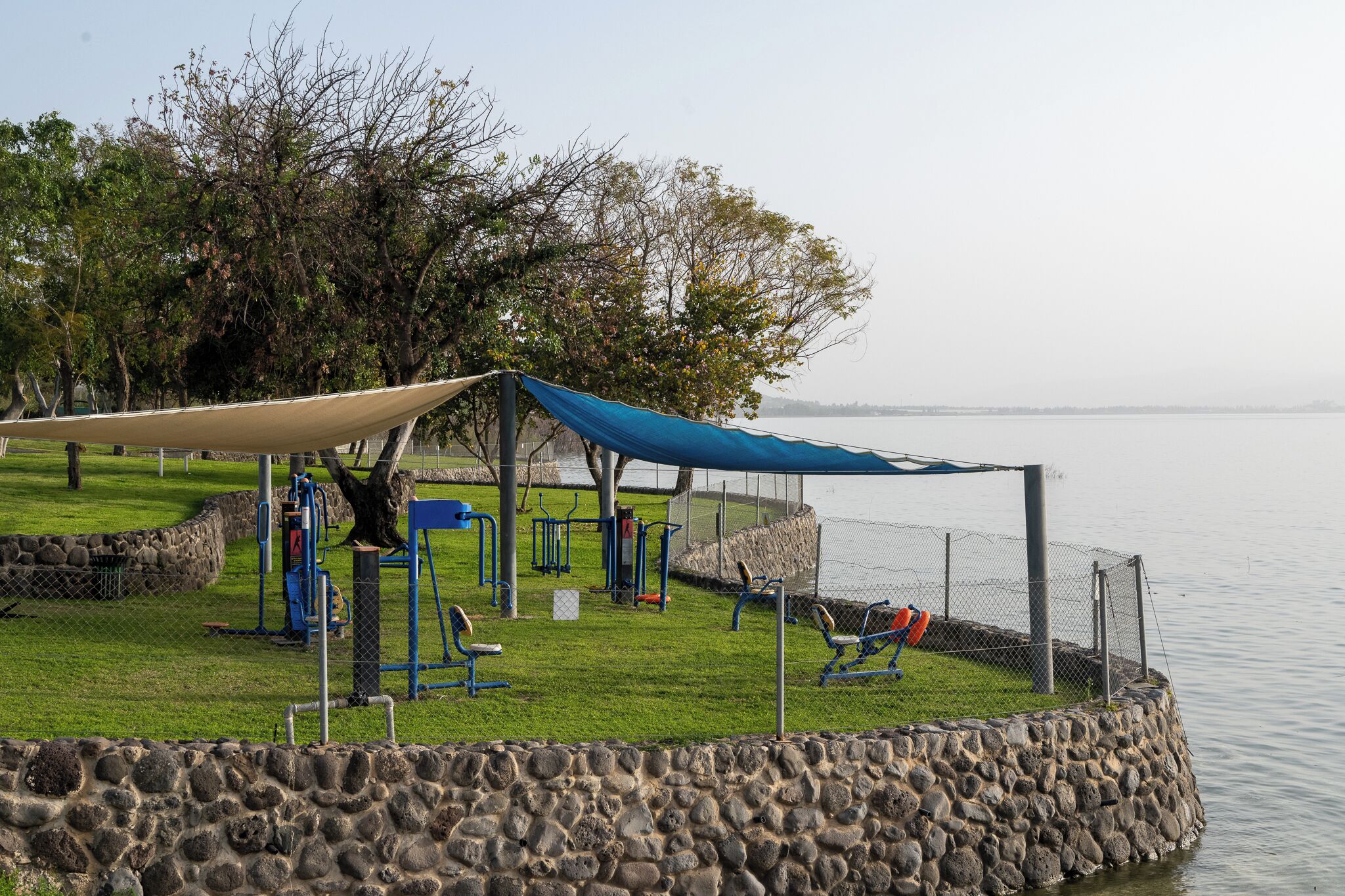Um parque com equipamentos de exercício, área coberta e um muro de pedra à beira da água.