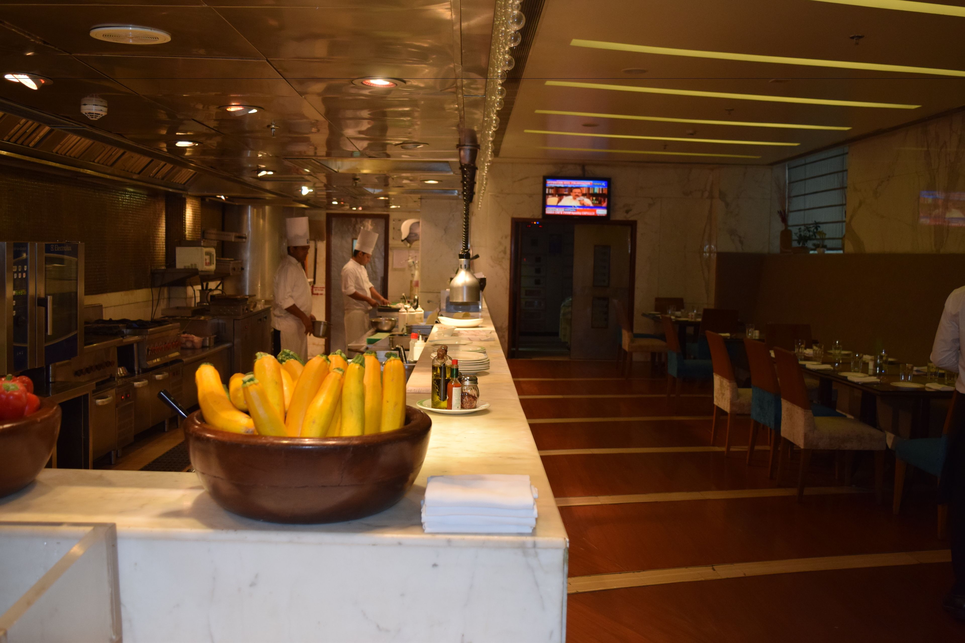 A restaurant with a long counter, a chef preparing food, and a television screen in the background.