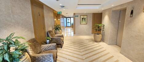 A hotel lobby with a herringbone patterned floor, a reception desk, and several seating areas with potted plants.