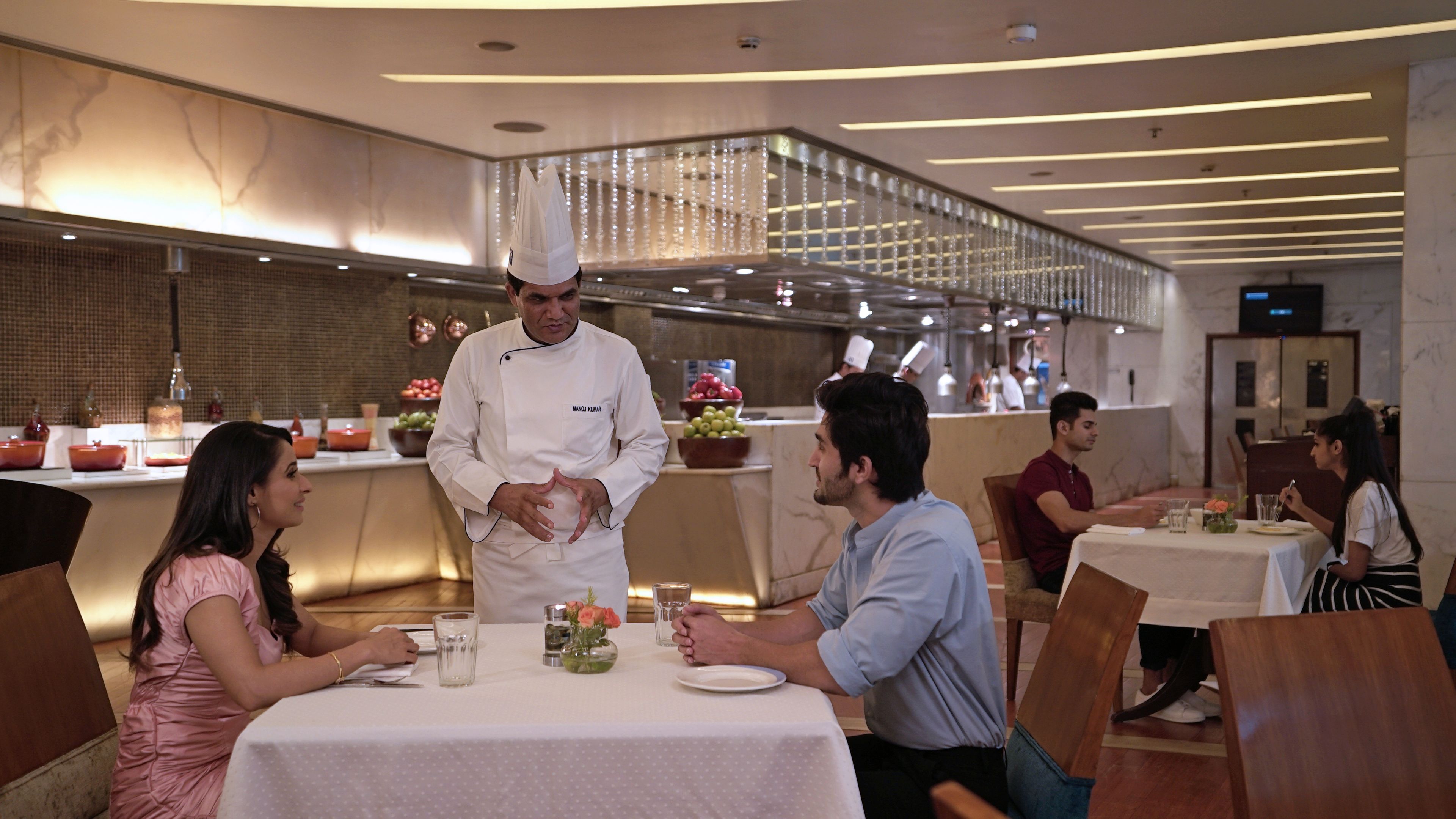 Interior d'un restaurant amb un xef al costat del taulell, taules amb clients asseguts i un disseny de sostre modern.