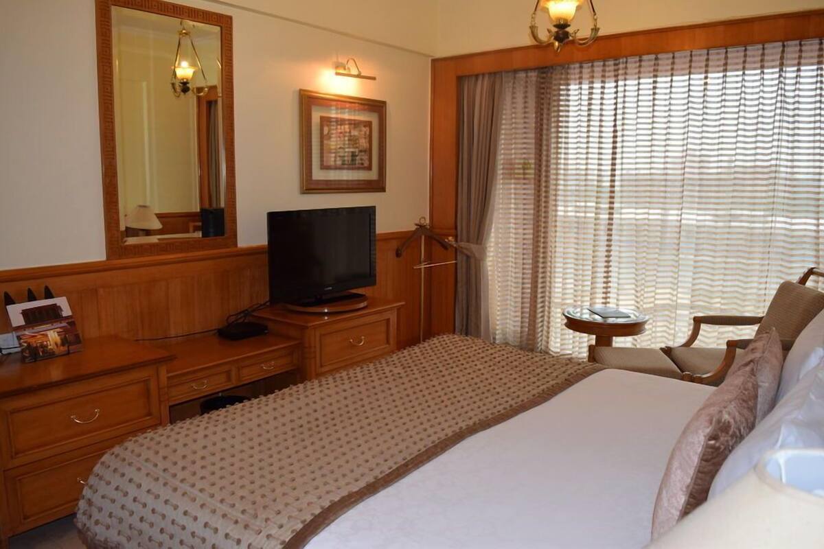 A hotel room with a bed, a television, a desk with drawers, a mirror, a framed picture, a window with curtains, and a small table with a lamp.