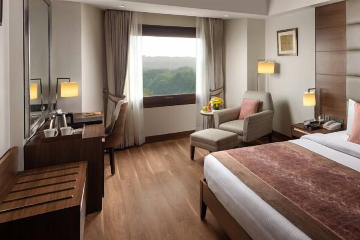 Habitación de hotel con una cama grande, un escritorio con silla, una mesita de noche con lámpara y una ventana con vistas a zonas verdes.
