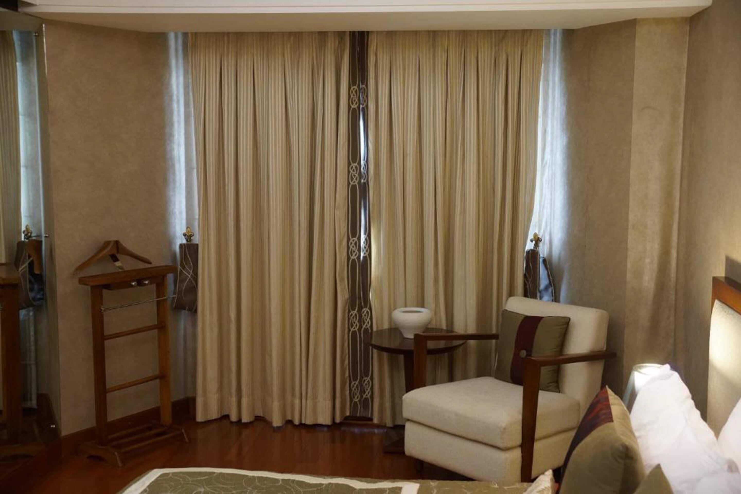 Una habitación de hotel con un ventanal grande, una silla, una mesita pequeña y un perchero.