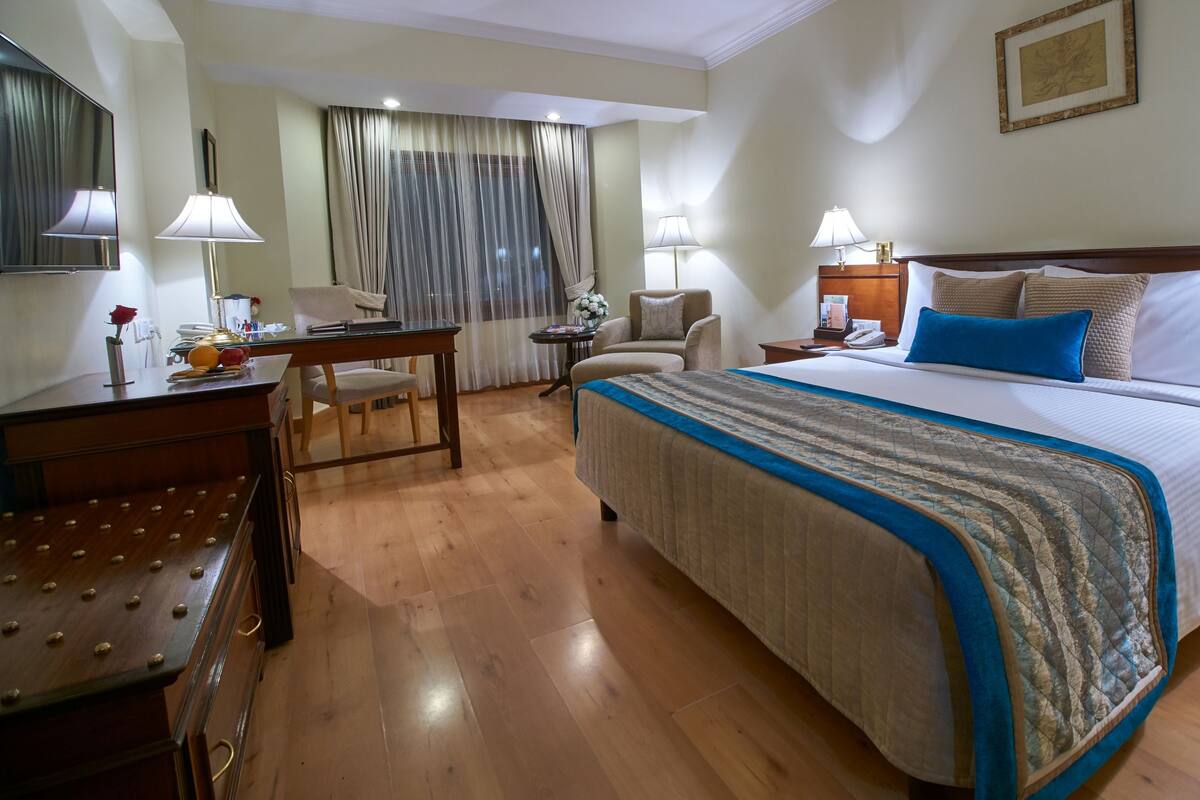 Habitación de hotel con una cama grande, un escritorio con lámpara, una silla y un ventanal con cortinas.