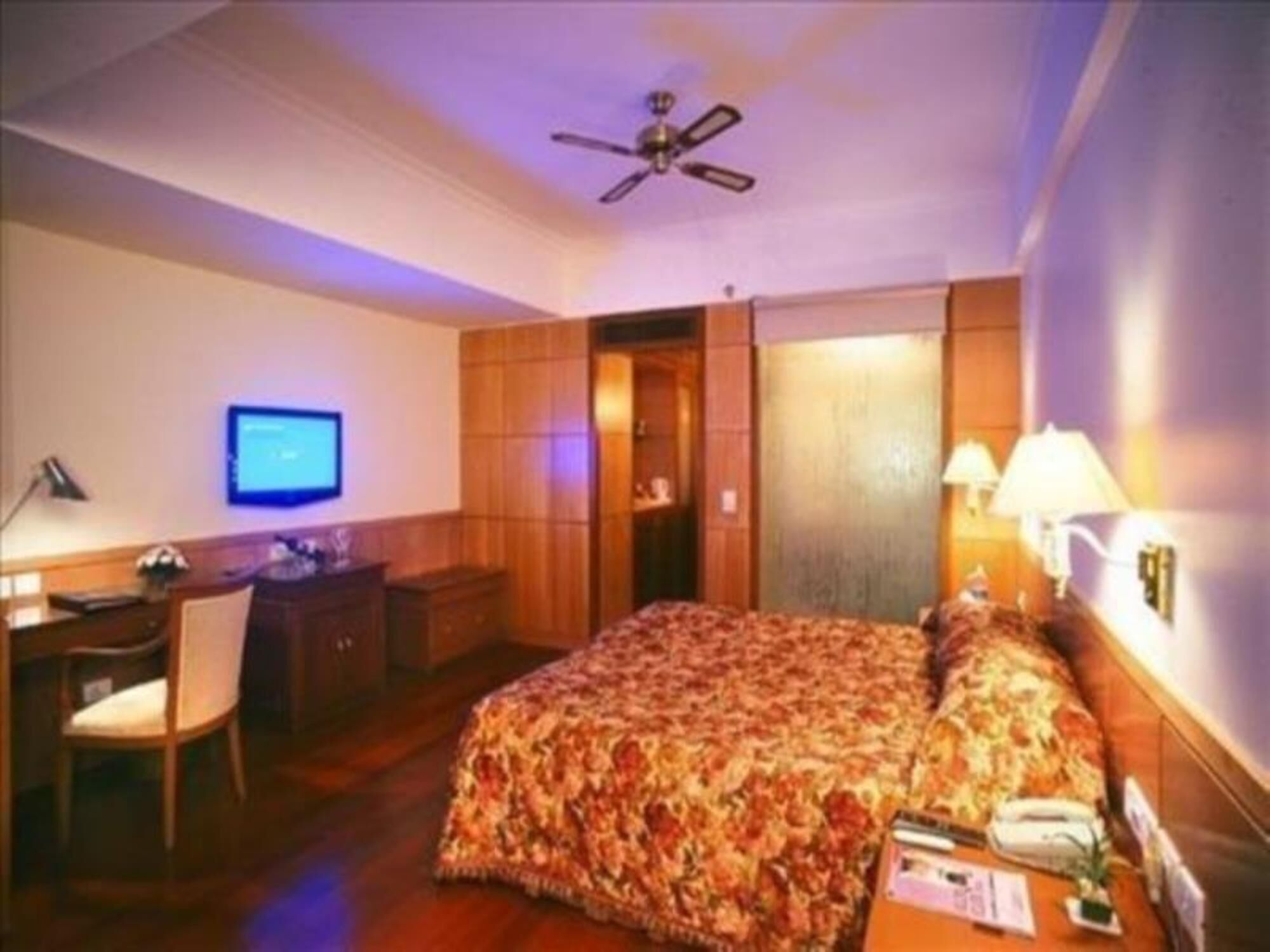 Una habitación de hotel con cama, escritorio, silla, televisor y ventilador de techo.