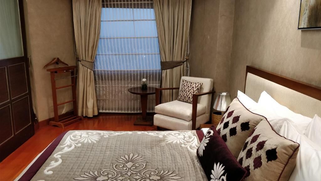 A hotel room with a bed, a chair, a small table, and a view of the outdoors through a window with curtains.