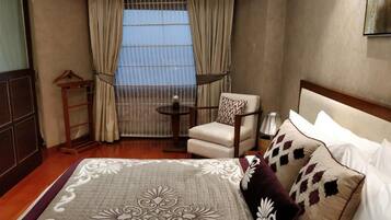 A hotel room with a bed, a chair, a small table, and a view of the outdoors through a window with curtains.
