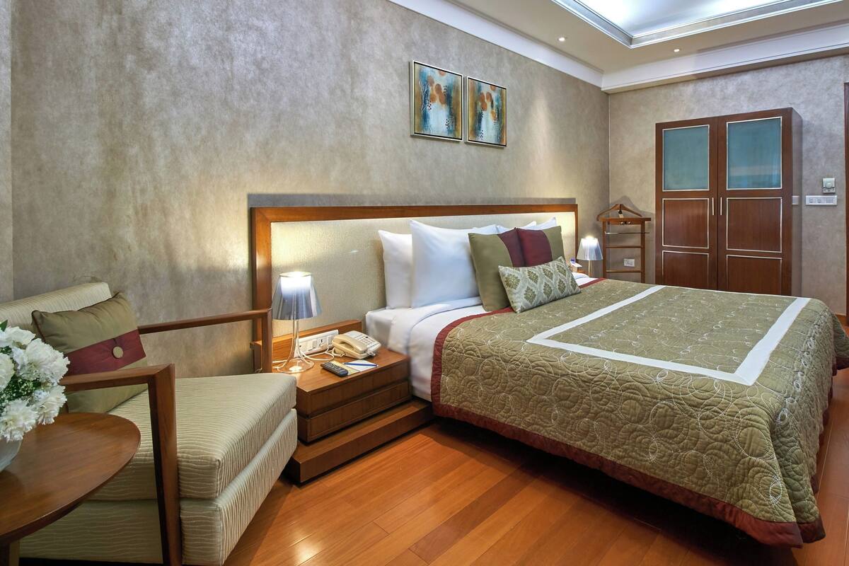 Habitación de hotel con una cama grande, una mesita de noche con lámpara, una silla, una pequeña mesa con un florero y un armario.