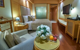 A hotel room with a bed, a sofa, a small table with a vase of flowers and fruit, a TV, and a wooden cabinet.