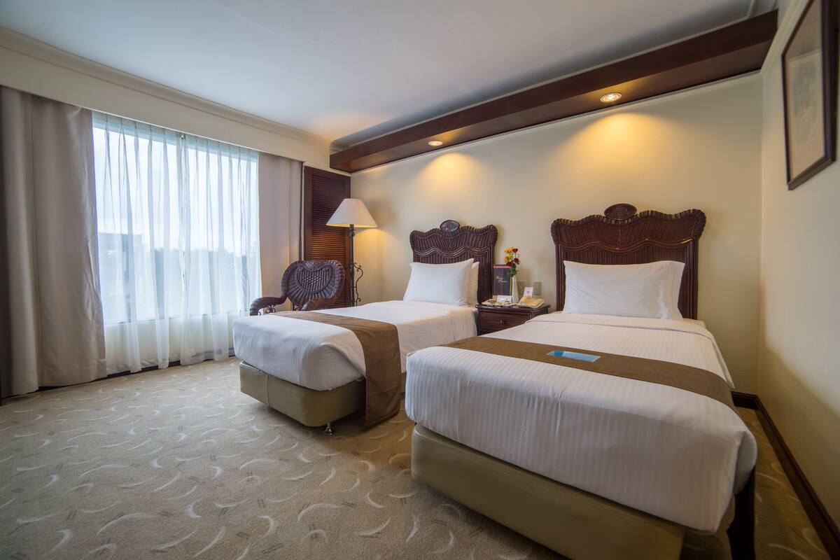 Habitación de hotel con dos camas, una mesita de noche, una lámpara y una ventana con cortinas.