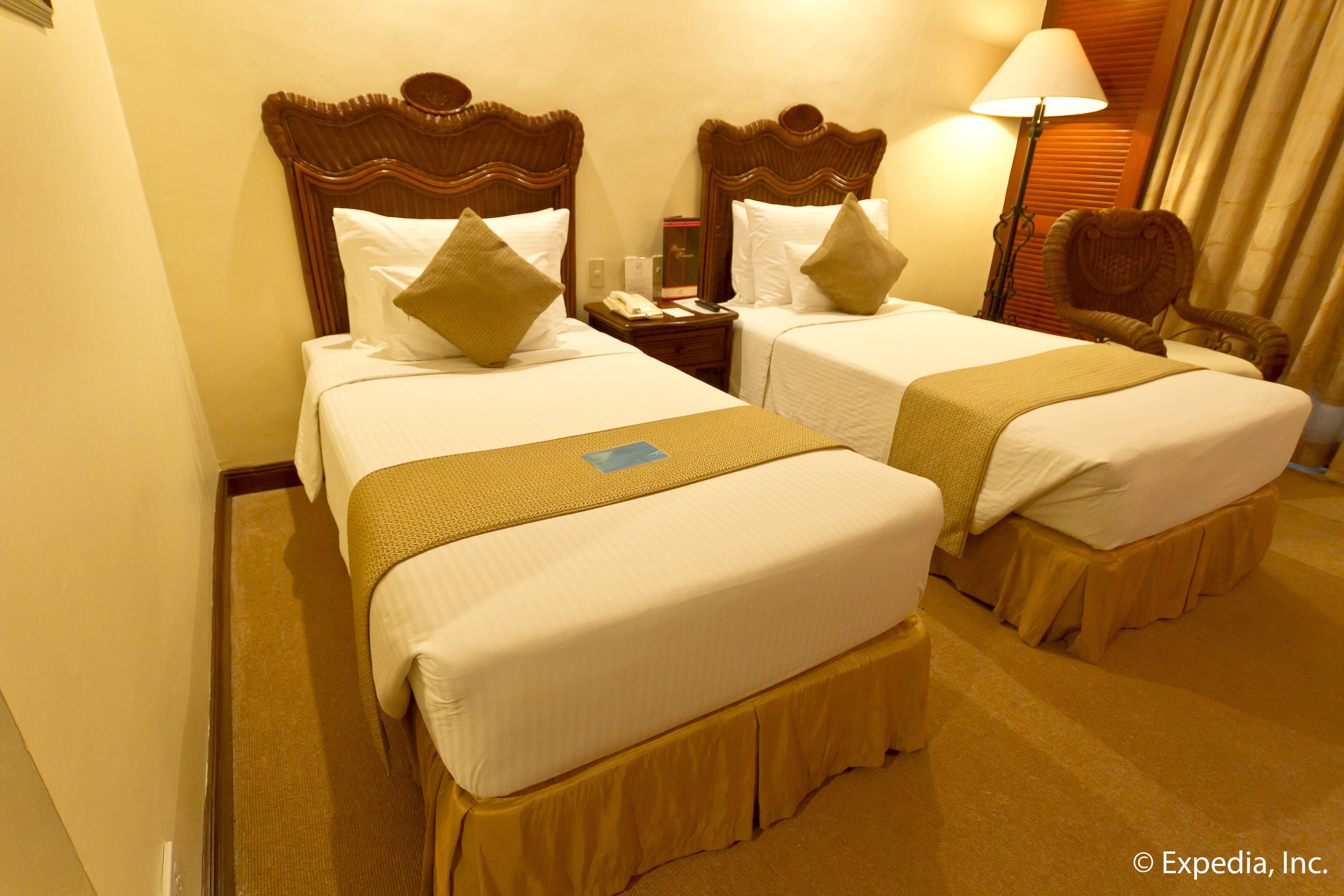 A hotel room with two beds, each with a wooden headboard, white linens, and a brown skirt. There is a nightstand with a lamp and a telephone between the beds.