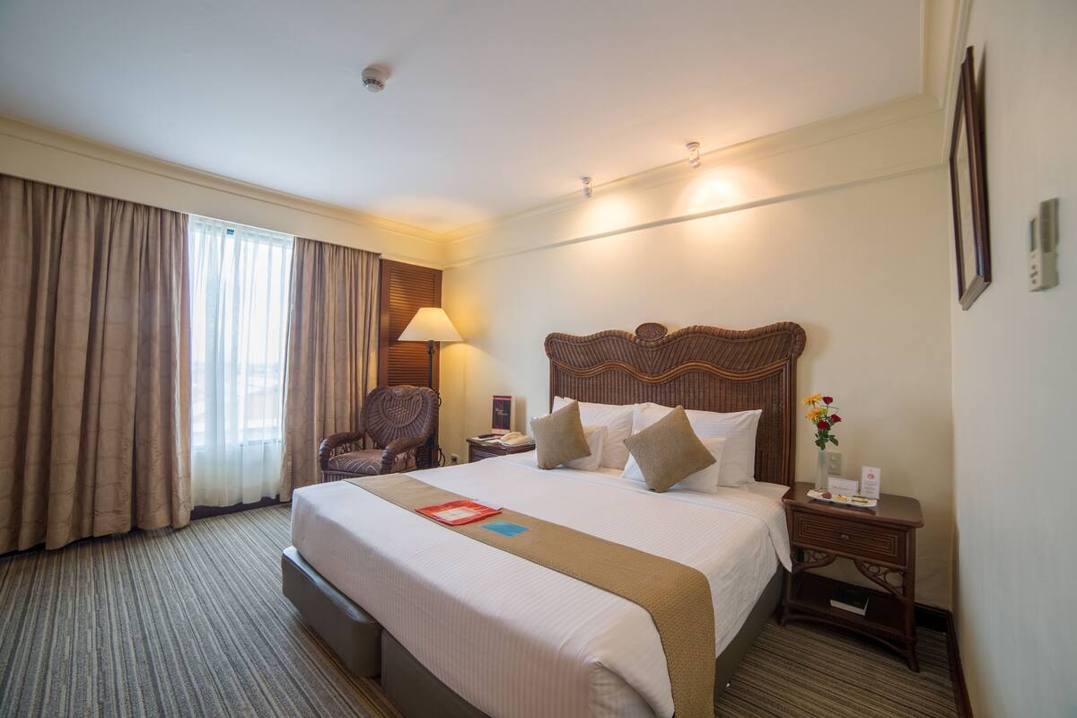 Una habitación de hotel con una cama grande, cabecera de madera, mesita de noche con lámpara, una silla y una ventana con cortinas.