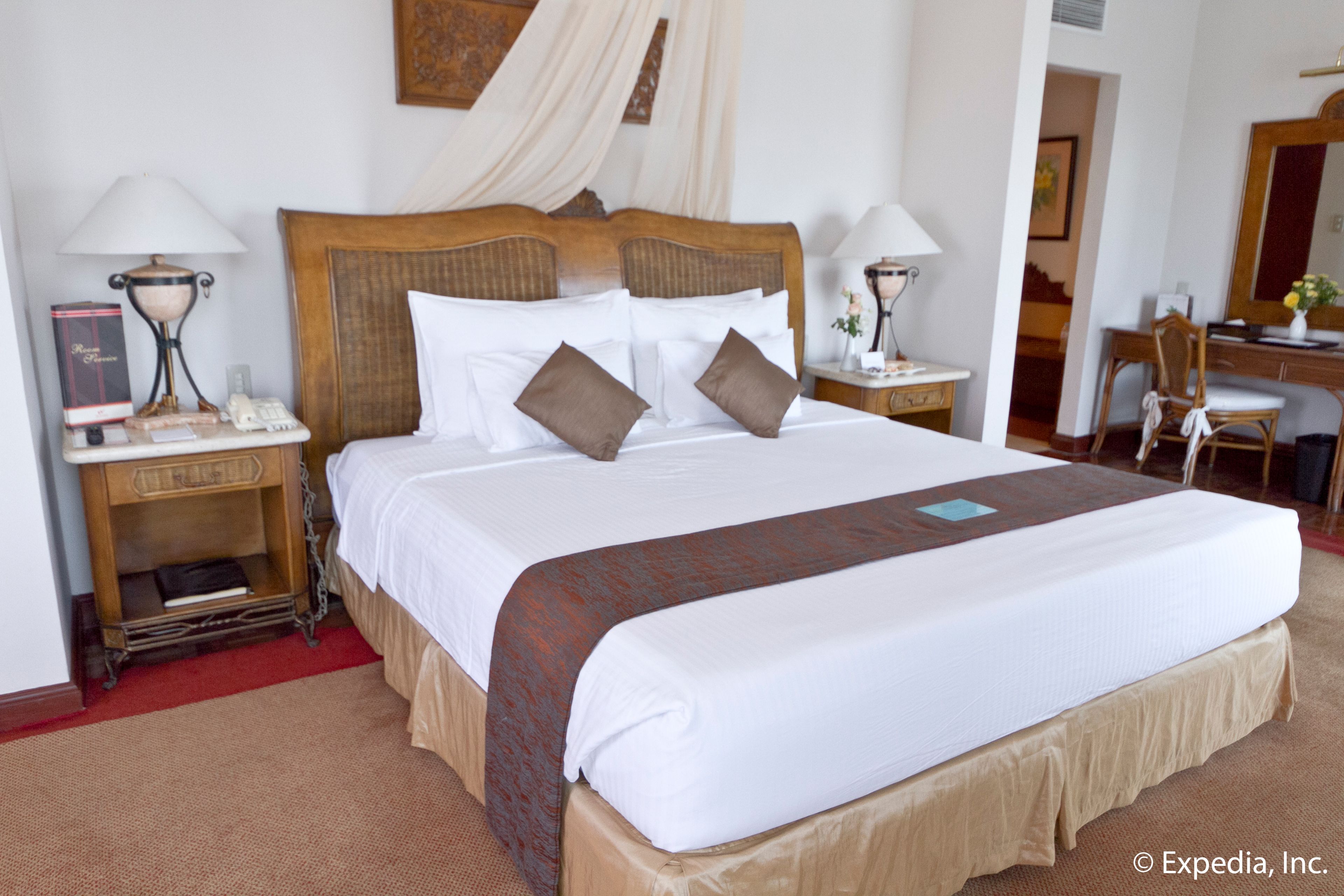 Una cama impecablemente tendida con sábanas blancas y un cojín marrón decorativo, un cabecero de madera y dos mesitas de noche con lámparas y un libro.