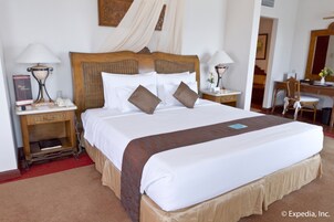 Una cama impecablemente tendida con sábanas blancas y un cojín marrón decorativo, un cabecero de madera y dos mesitas de noche con lámparas y un libro.