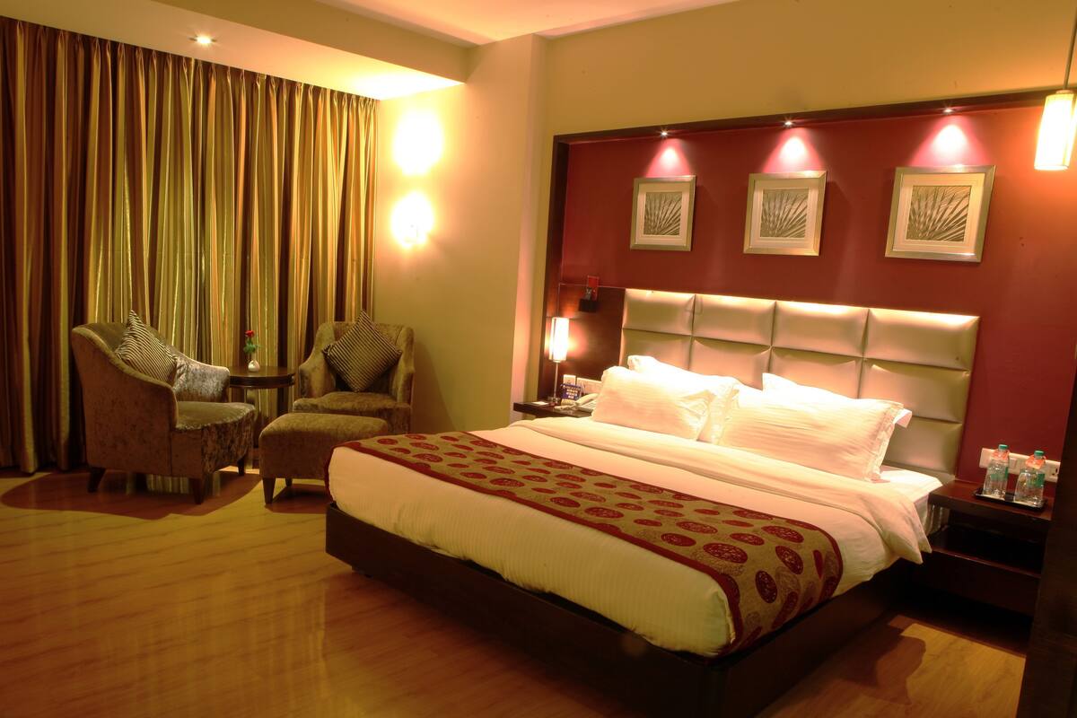 Habitación de hotel con una cama grande, dos sillones, una mesita y una mesita de noche con lámpara y botellas de agua.