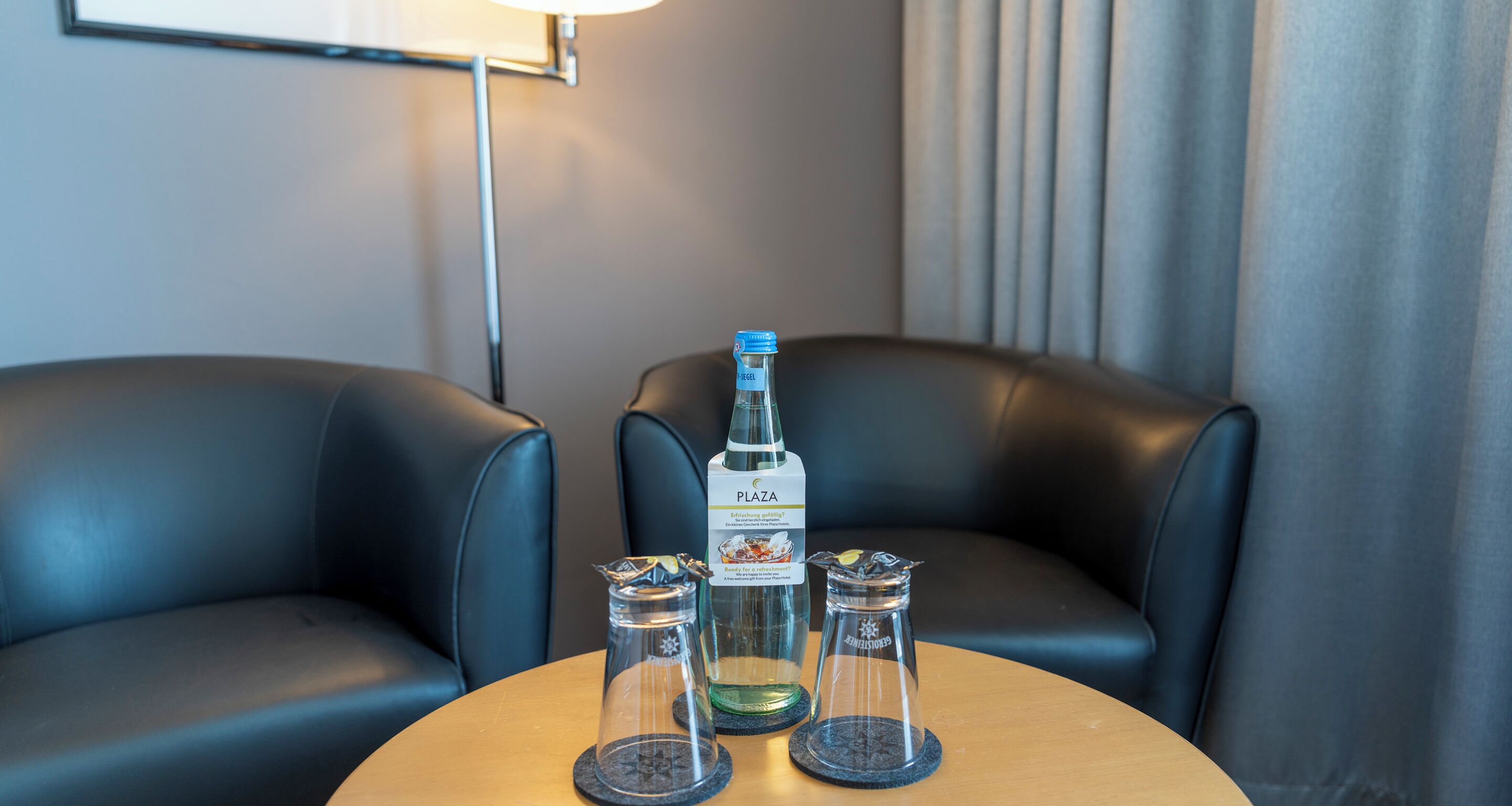A table with a bottle of PLAZA and two glass carafes with a spoon, set between two black leather chairs.