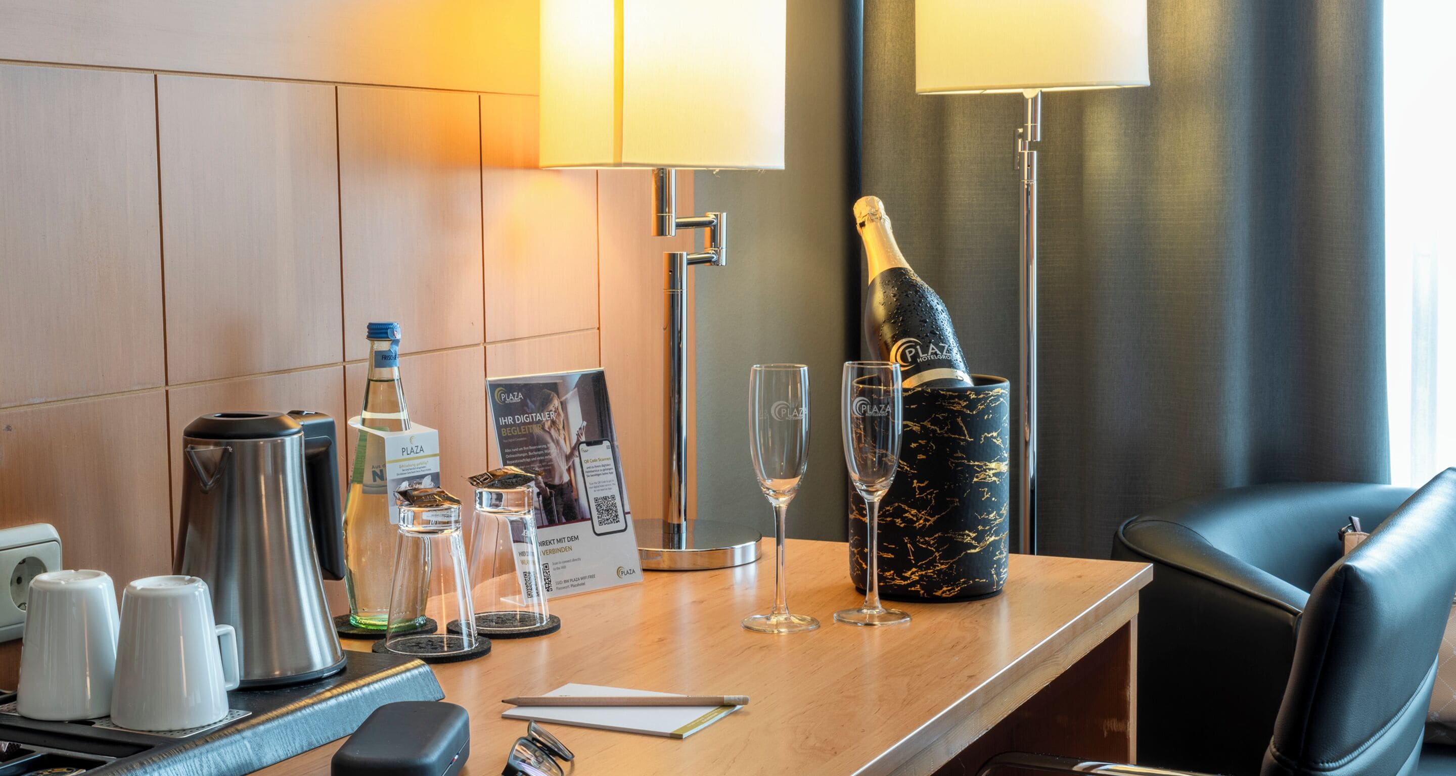 Een bureau in een hotelkamer met een koffiezetapparaat, twee bekers, een fles champagne, glazen, een lamp en een stoel.
