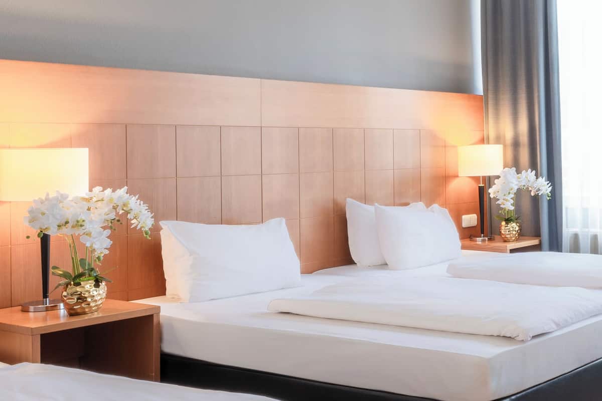 Ein Hotelzimmer mit einem großen Bett, zwei Nachttischen mit Lampen und Blick ins Freie durch ein Fenster mit Vorhängen.