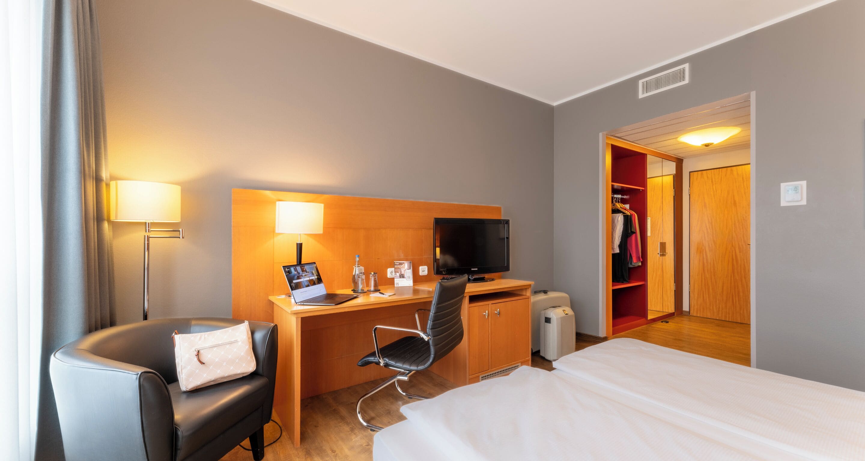 Una camera d'albergo con un letto, una scrivania con un portatile, una sedia, una televisione e un armadio.