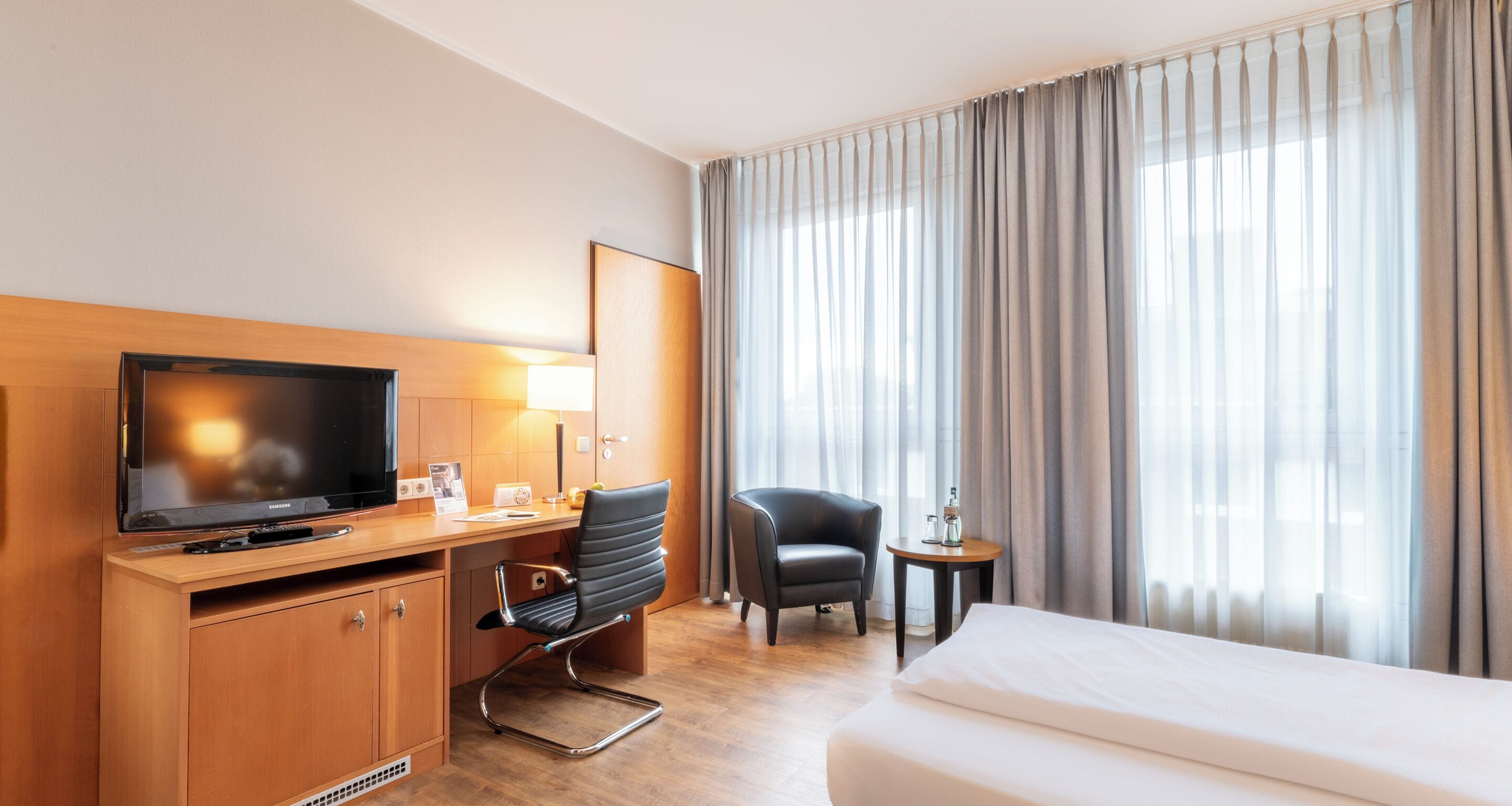 A hotel room with a bed, a desk with a television, a chair, and a small table.