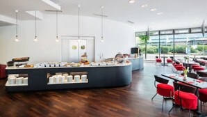 A modern dining area with a central buffet station, multiple seating arrangements, and large windows offering a view of the outdoors. - Penck Hotel Dresden (Dresden)