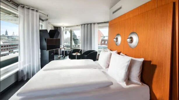 A hotel room with a large bed, a wooden accent wall, a TV, and a view of a cityscape. - Penck Hotel Dresden (Dresden)