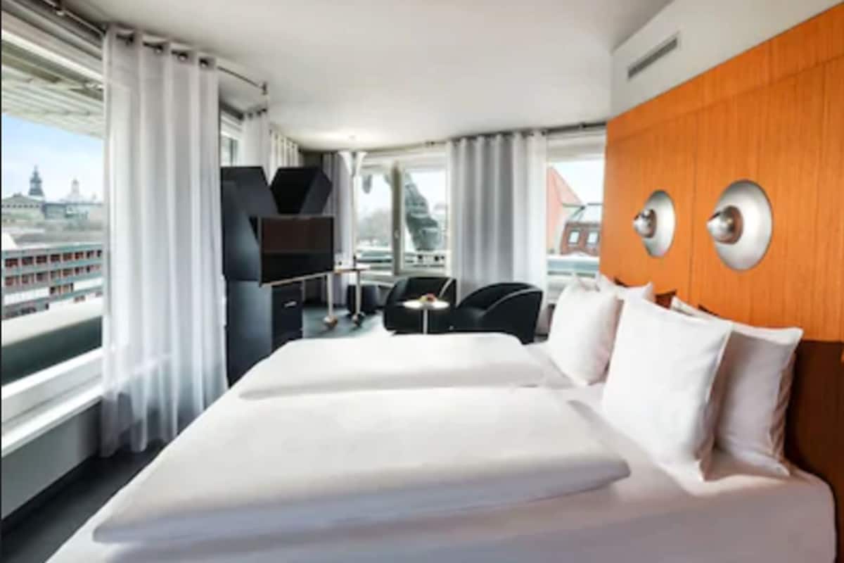 Habitación de hotel con una cama grande, un muro decorativo de madera, un televisor y vistas a un paisaje urbano.
