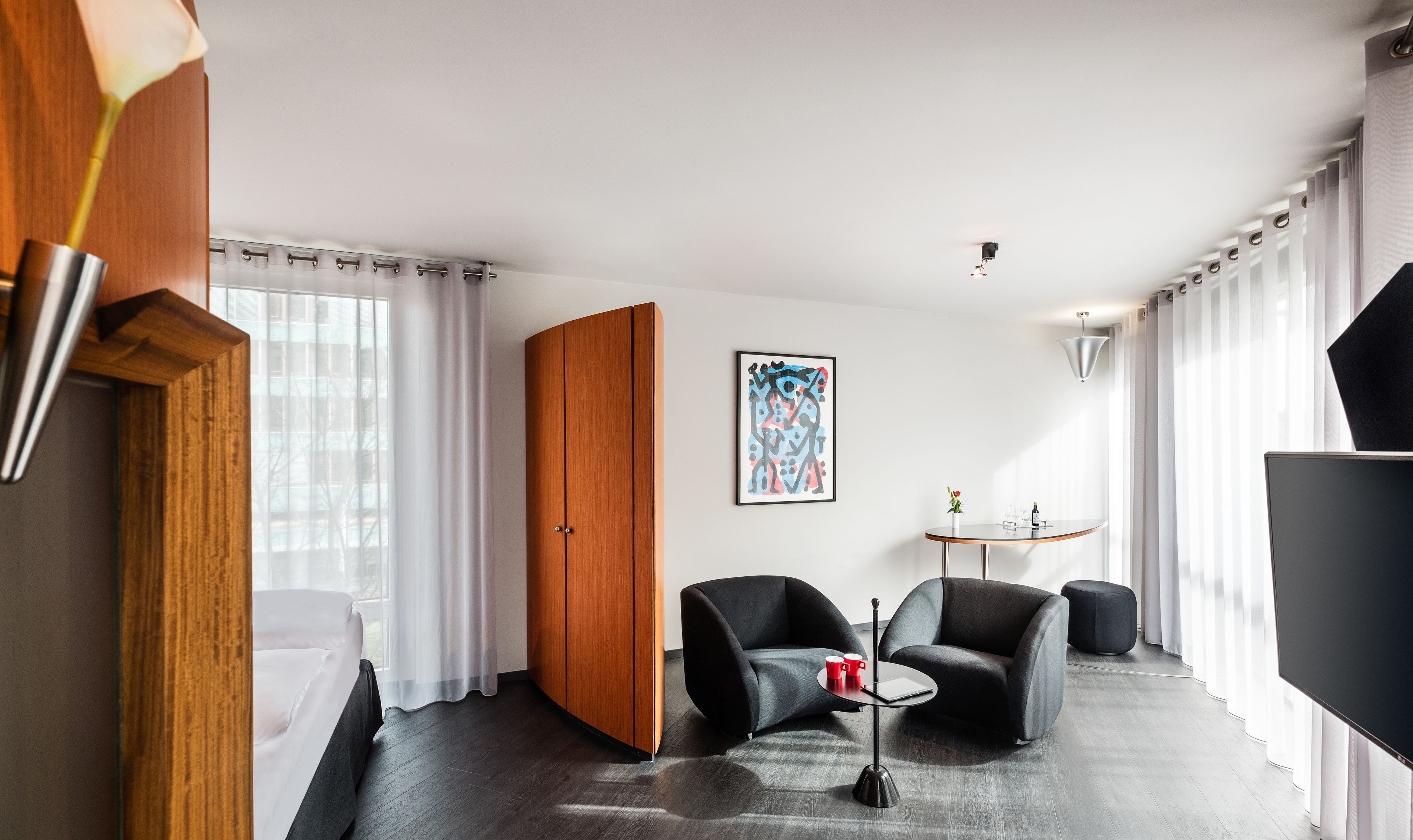Een moderne hotelkamer met een bed, twee fauteuils, een kleine tafel, een televisie en een schilderij aan de muur.