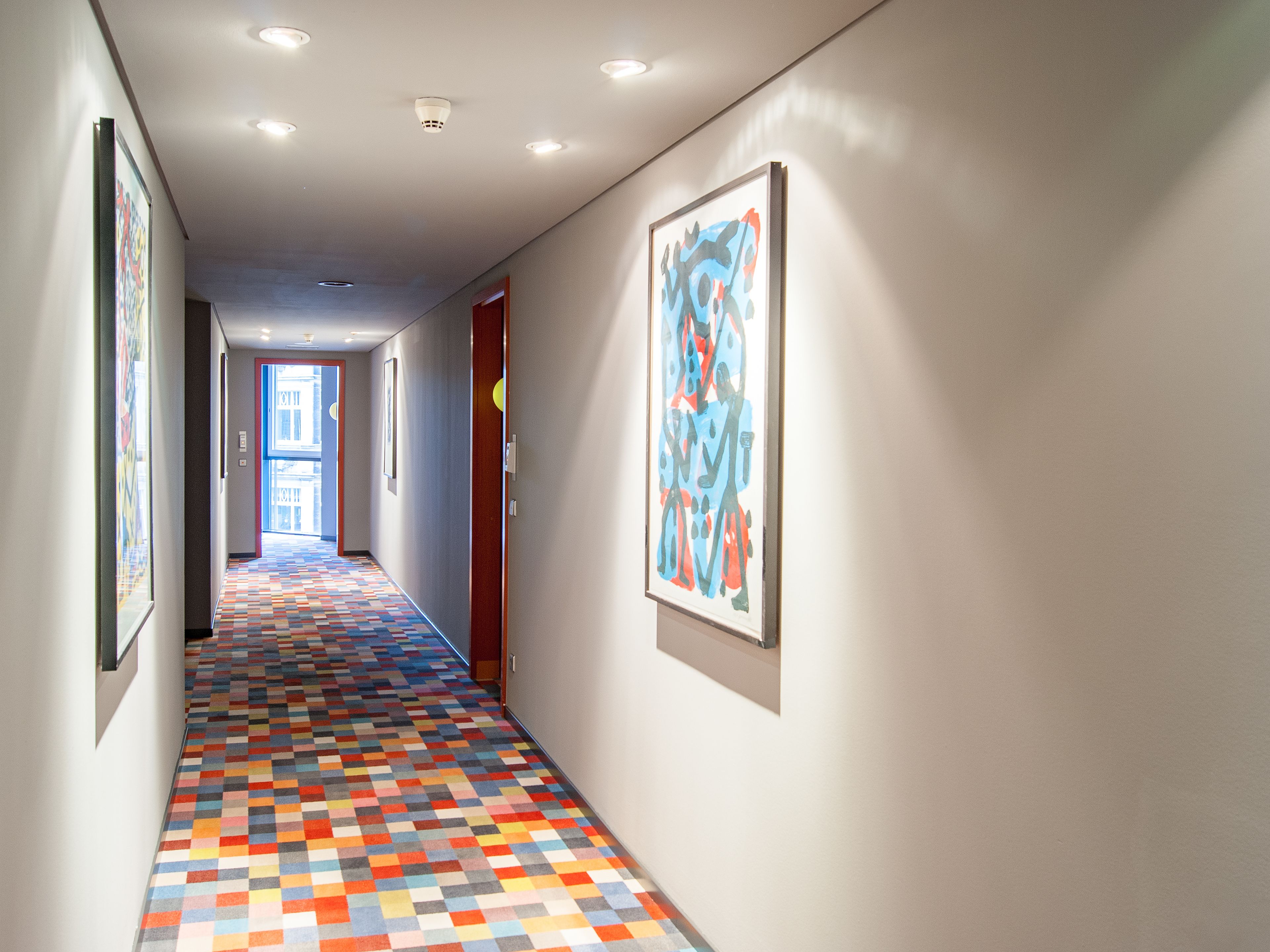 Un pasillo con piso a cuadros de colores, obras de arte enmarcadas en las paredes e iluminación empotrada en el techo.